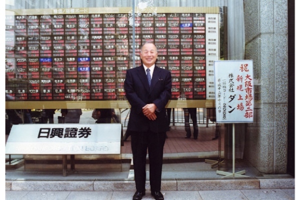 2000年の大証二部上場時、株価ボードの前に立つ越智直正氏（提供：タビオ）